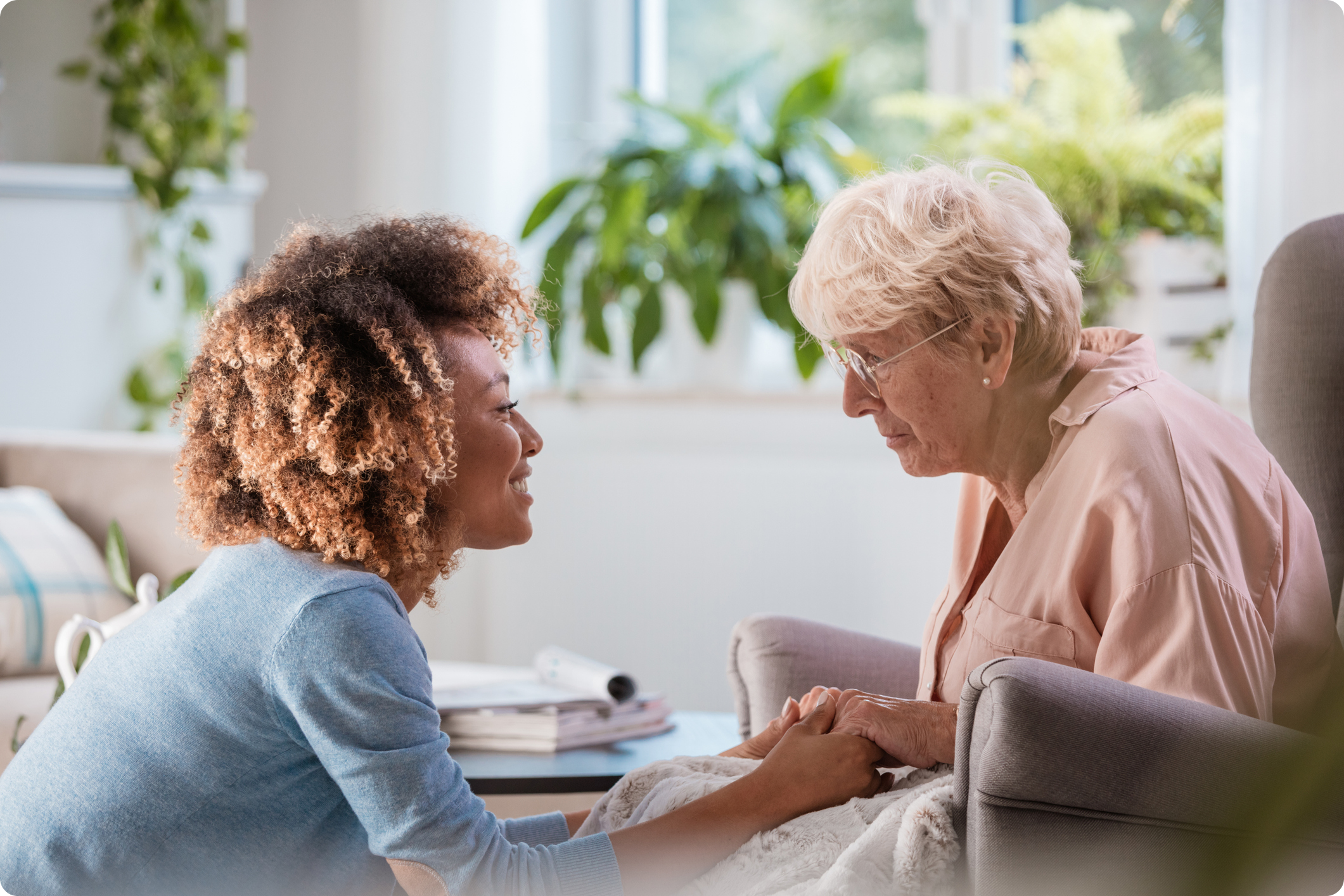 A caregiver interacts with an elderly woman, holding hands in a bright, plant-filled room promoting comfort and connection.