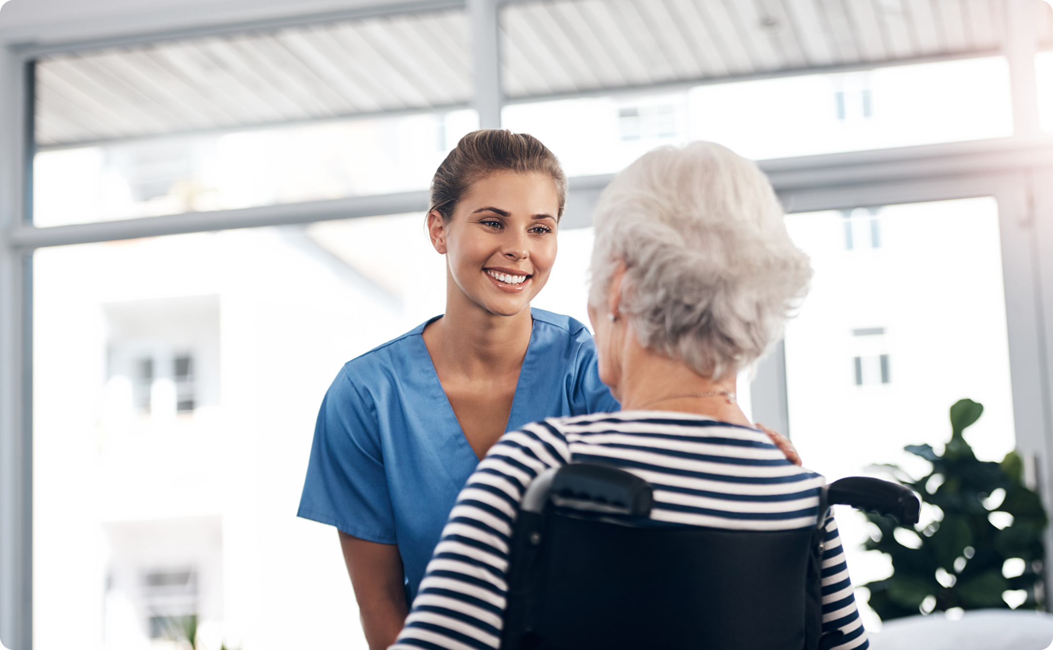 Healthcare professional in blue scrubs smiling while engaging with an elderly woman in a striped shirt seated in a wheelchair.