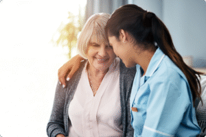 A caregiver embraces an elderly woman, both smiling warmly in a softly lit room.