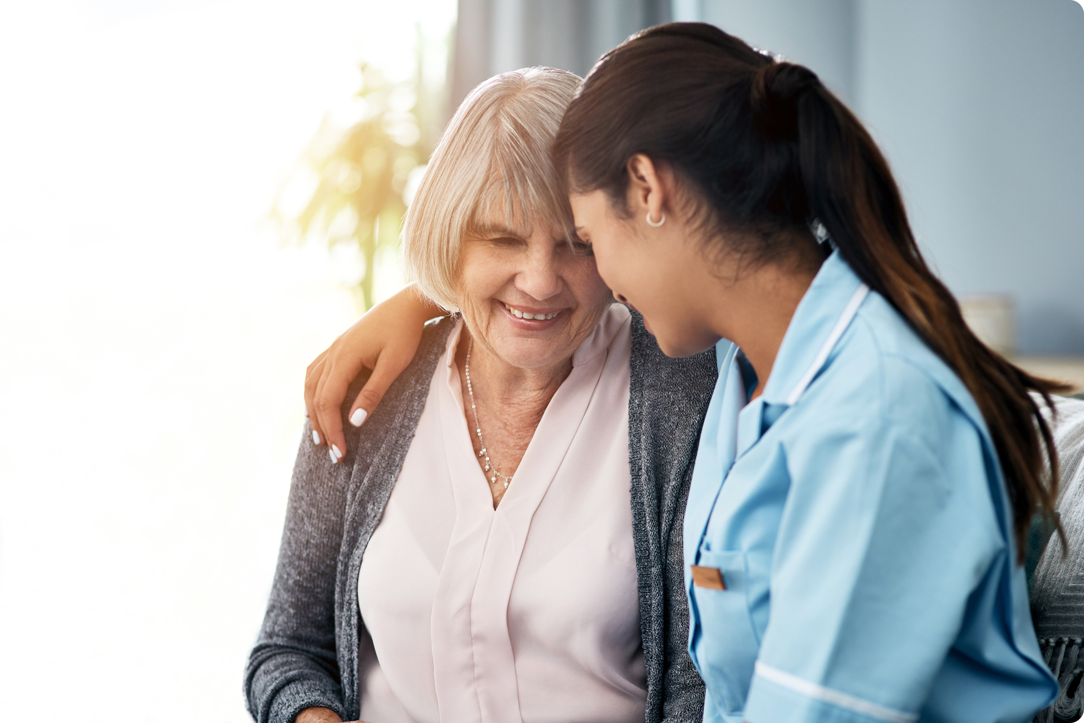 A caregiver embraces an elderly woman, both smiling warmly in a softly lit room.