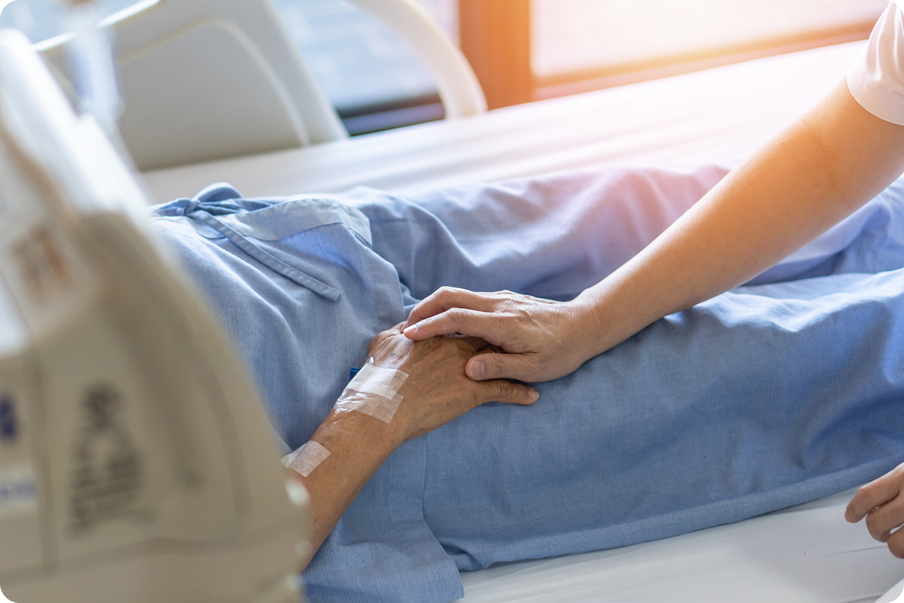 A nurse gently holds the hand of a patient in a hospital bed, surrounded by medical equipment and natural light.