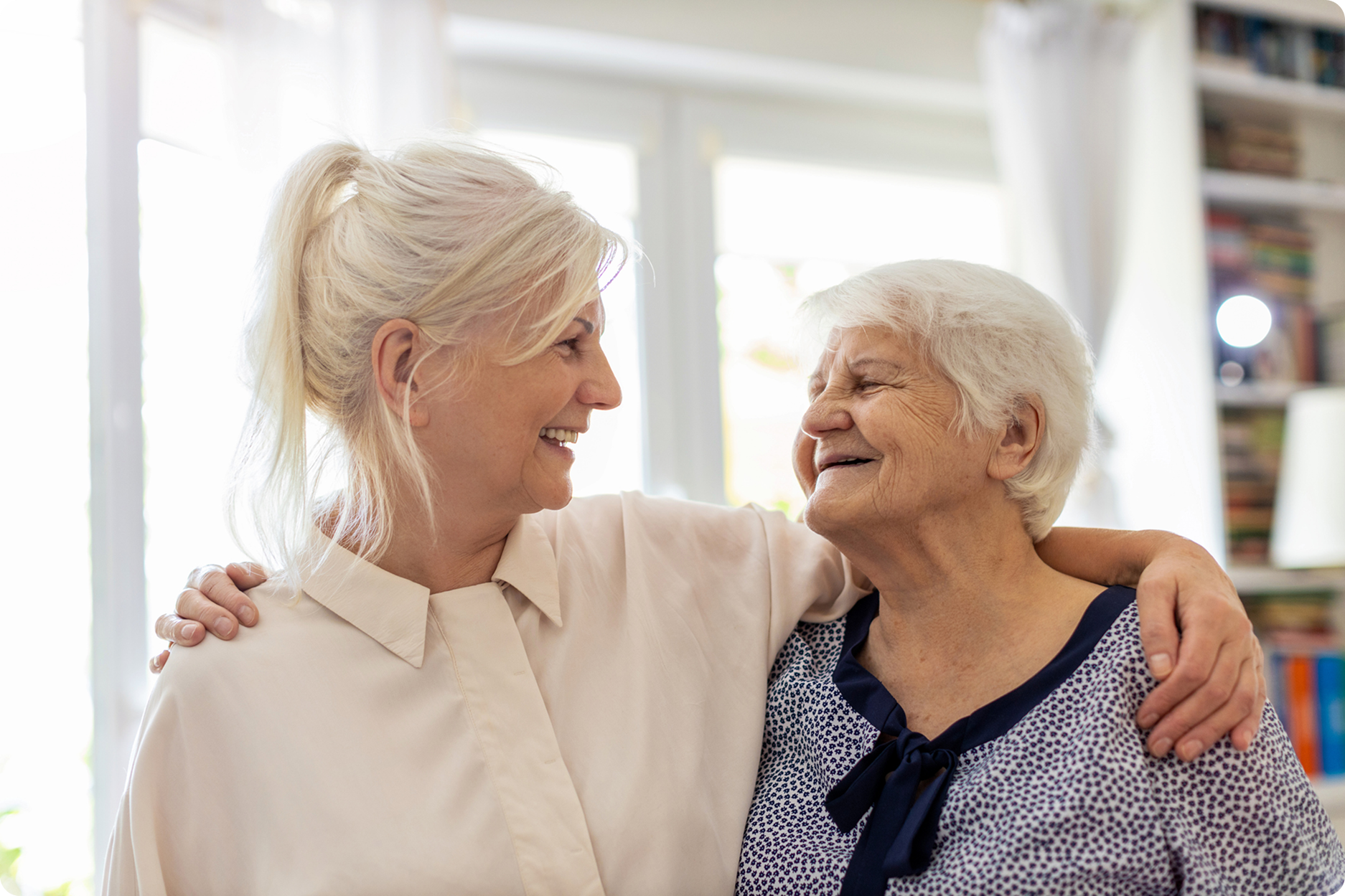 Two women smiling and embracing in a bright, cozy living room filled with natural light and bookshelves in the background.