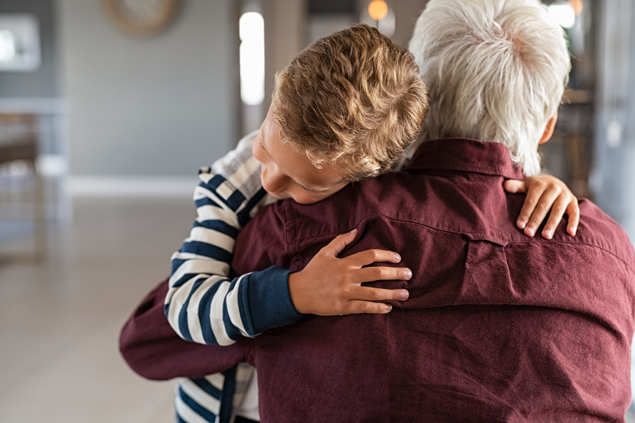 A young boy hugs an older adult from behind, both smiling, in a warmly lit indoor setting.