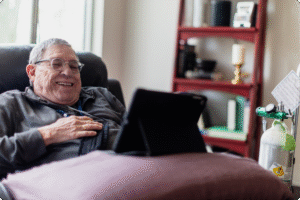 Senior man smiling while using a tablet, sitting on a comfortable chair with an oxygen tank and bookshelf in the background.