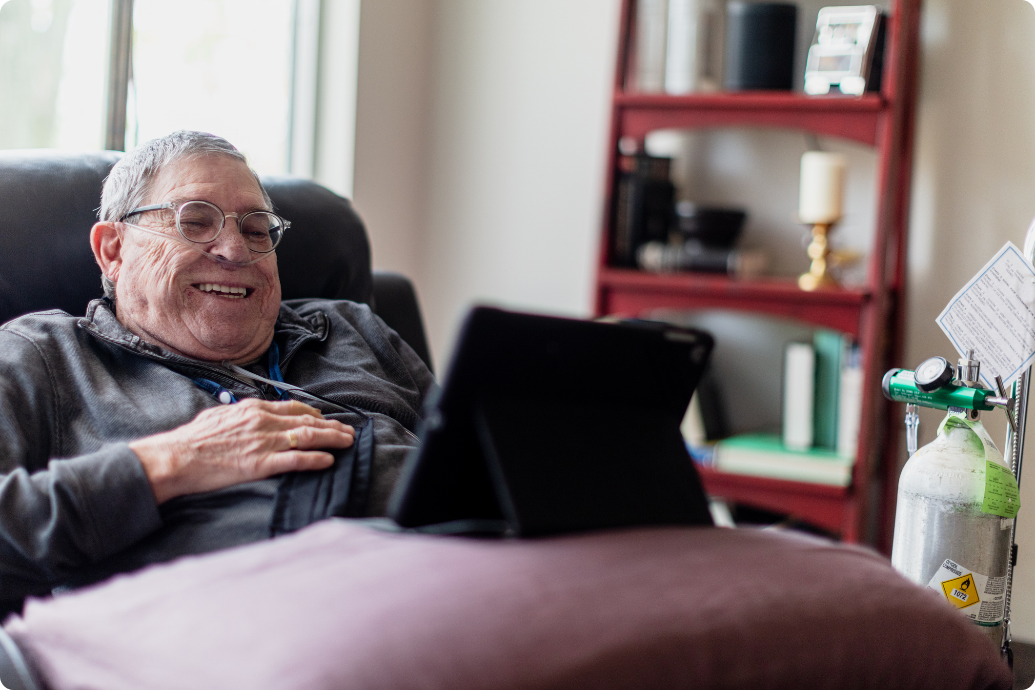 Senior man smiling while using a tablet, sitting on a comfortable chair with an oxygen tank and bookshelf in the background.