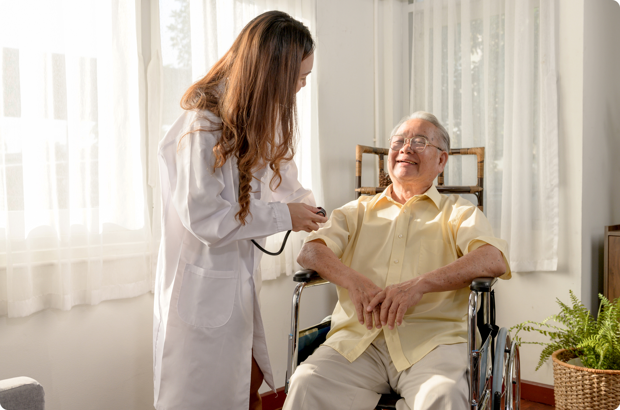 A healthcare professional in a white coat checks the blood pressure of a smiling elderly man in a wheelchair.