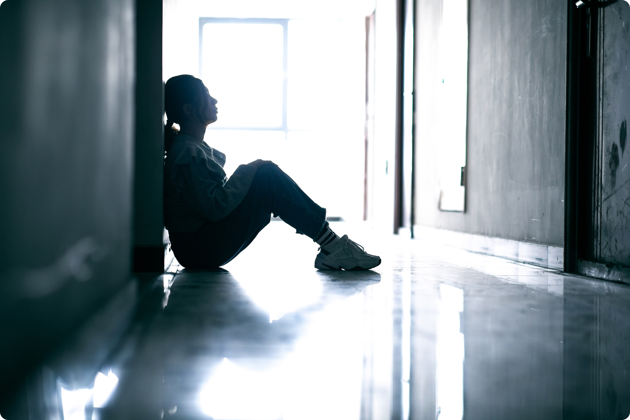 A young woman sits against a wall in a dim hallway, illuminated by light coming from a window at the end.