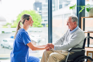 Healthcare professional and elderly patient in a wheelchair engage in conversation, seated by large windows in a bright, modern environment.