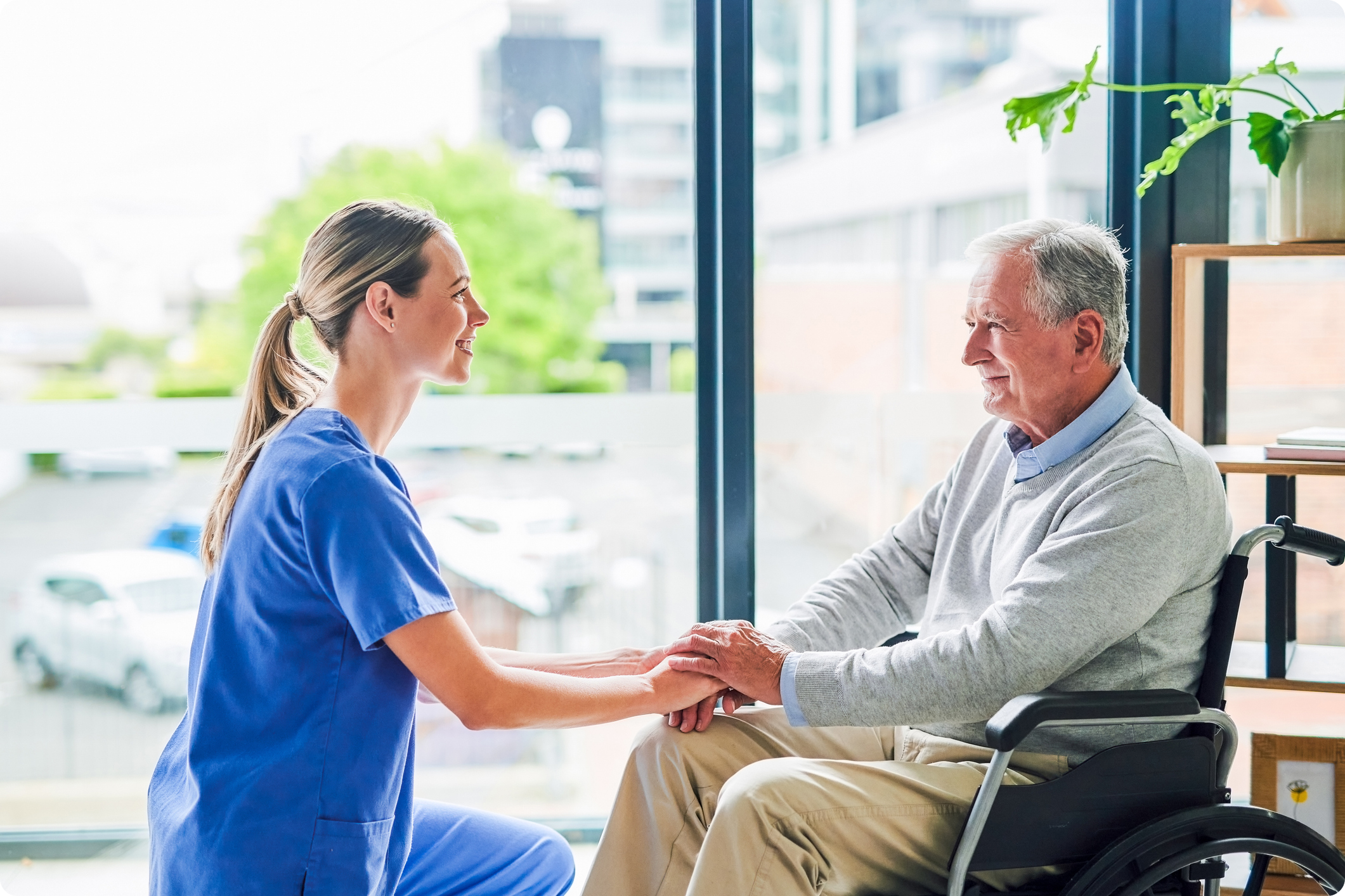 Healthcare professional and elderly patient in a wheelchair engage in conversation, seated by large windows in a bright, modern environment.