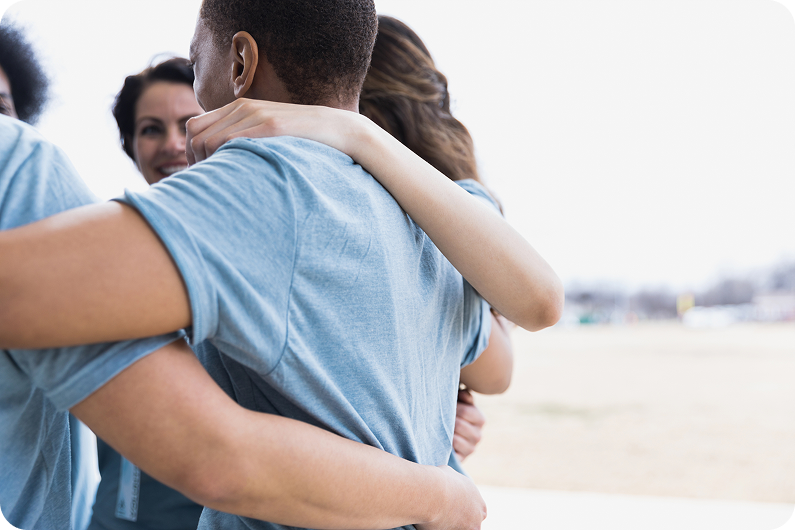 Group of individuals embracing in casual blue shirts, smiling and standing outdoors in a friendly gathering.