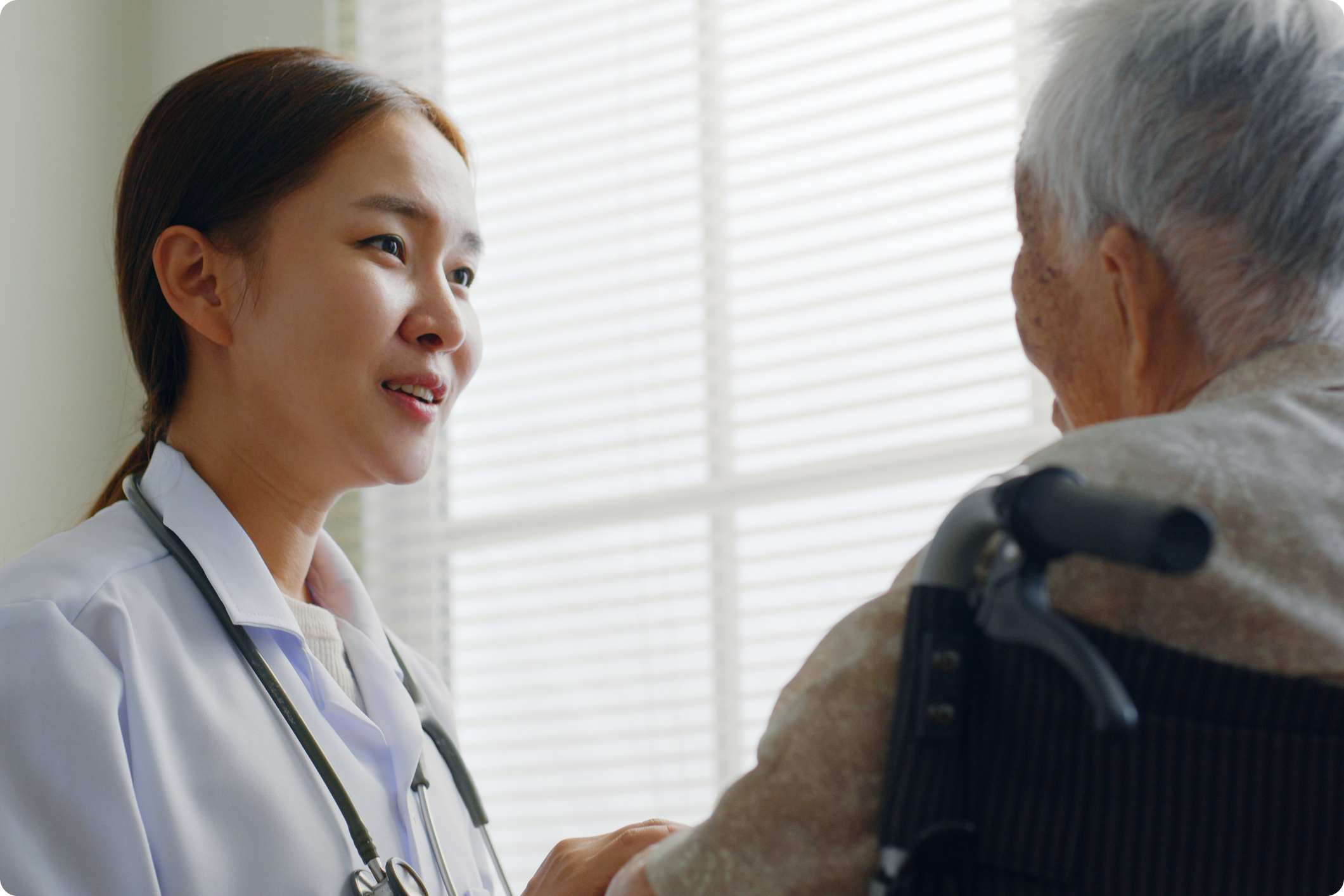 A healthcare professional engages with an elderly patient in a well-lit room with blinds.