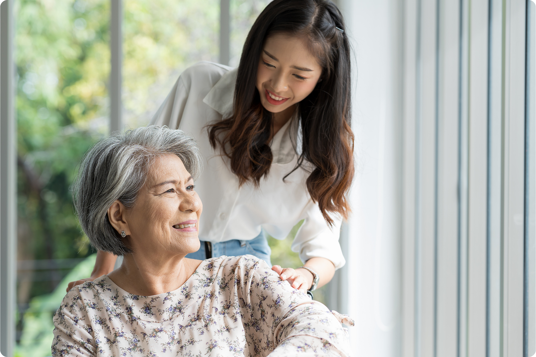 An older woman smiles while a younger woman gently touches her shoulder, set in a bright, sunlit environment.
