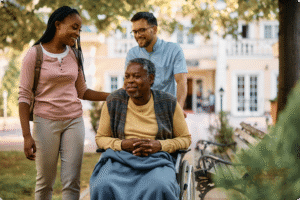 Two caregivers assist a smiling man in a wheelchair, surrounded by greenery and a sunny courtyard setting.