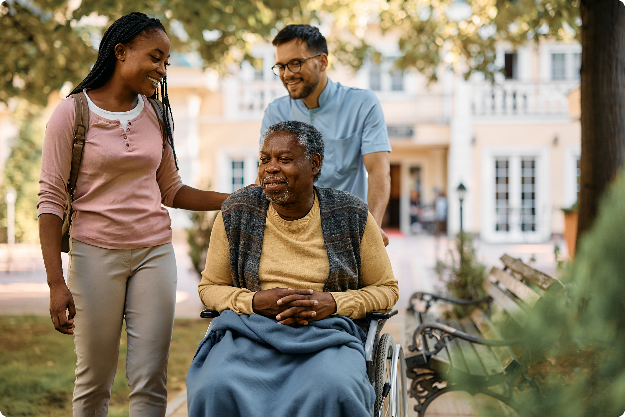 Two caregivers assist a smiling man in a wheelchair, surrounded by greenery and a sunny courtyard setting.
