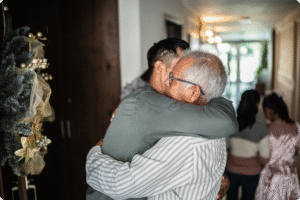 Two men embrace warmly in a welcoming hallway decorated for the holidays, with family members in the background.