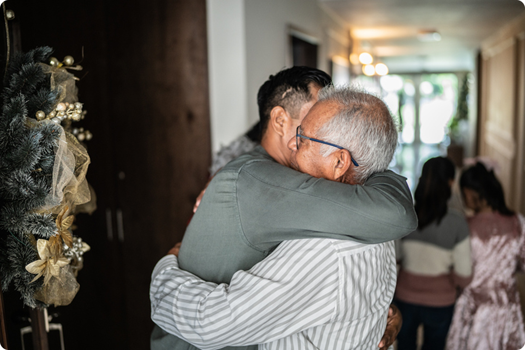 Two men embrace warmly in a welcoming hallway decorated for the holidays, with family members in the background.