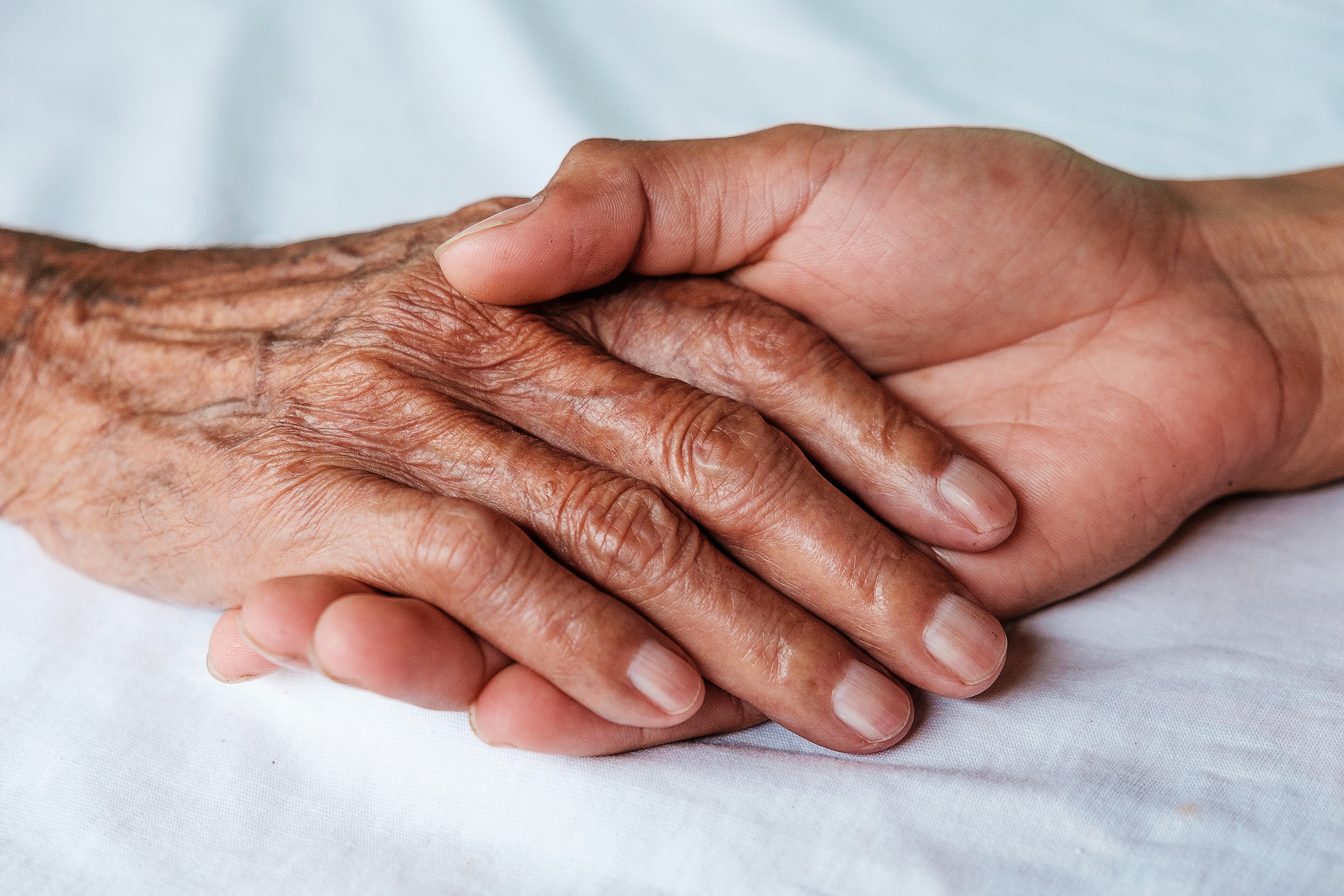 Two hands, one elderly and wrinkled, resting gently atop a younger hand on a white fabric surface.