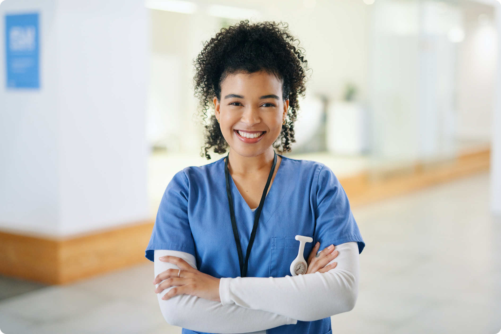 Smiling healthcare professional in a blue scrubs with arms crossed in a modern medical facility setting.