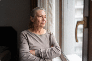 An elderly woman with grey hair stands by a window, looking thoughtfully outside, arms crossed in a cozy interior.