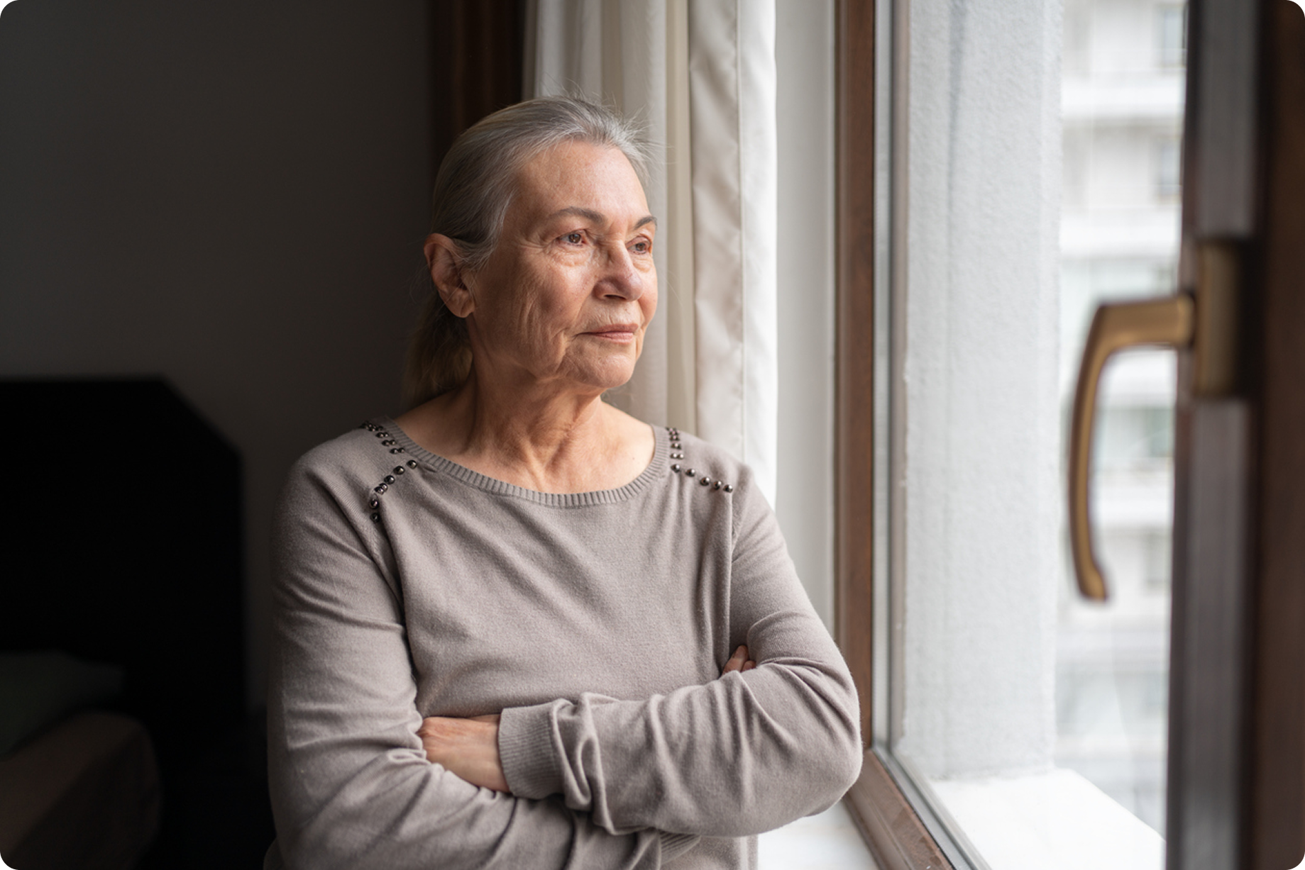 An elderly woman with grey hair stands by a window, looking thoughtfully outside, arms crossed in a cozy interior.