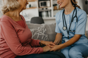 A healthcare professional engages in a compassionate conversation while holding the hands of an elderly patient in a home setting.