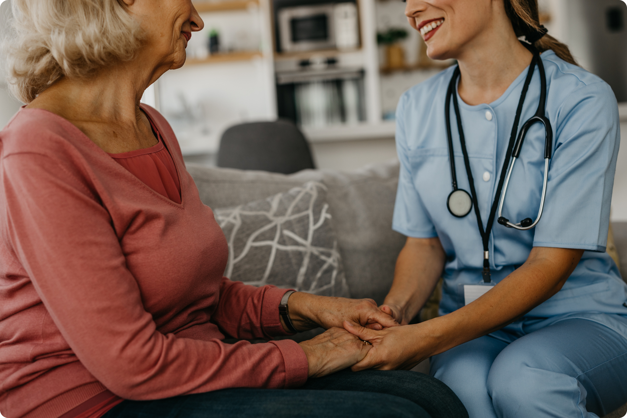 A healthcare professional engages in a compassionate conversation while holding the hands of an elderly patient in a home setting.