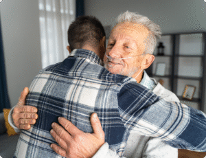 An elderly man with a nasal cannula embraces a younger man in a warmly lit, modern indoor setting.