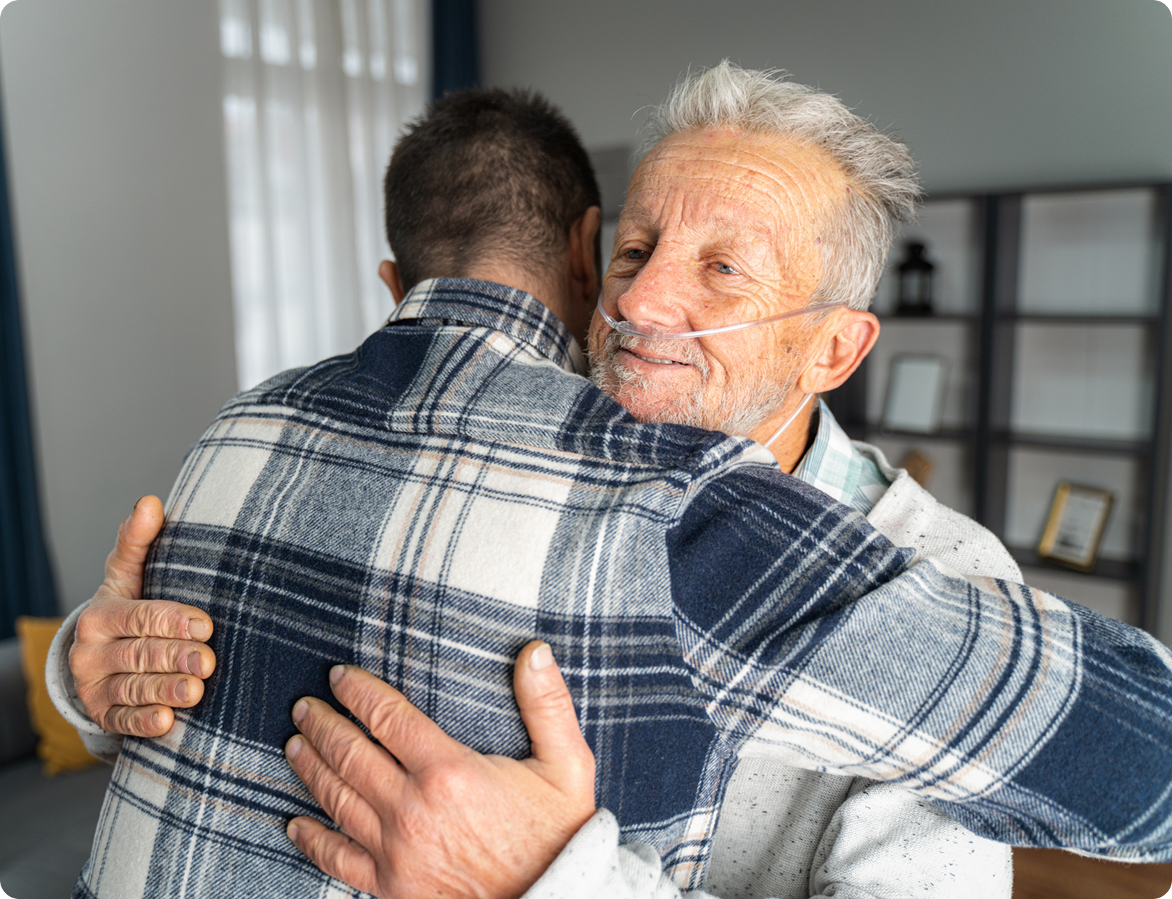 An elderly man with a nasal cannula embraces a younger man in a warmly lit, modern indoor setting.