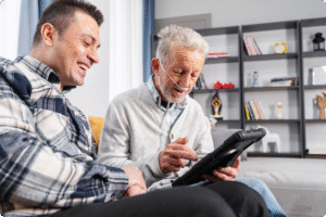 An older man with a nasal cannula smiles while using a tablet alongside a younger man in a cozy living room setting.