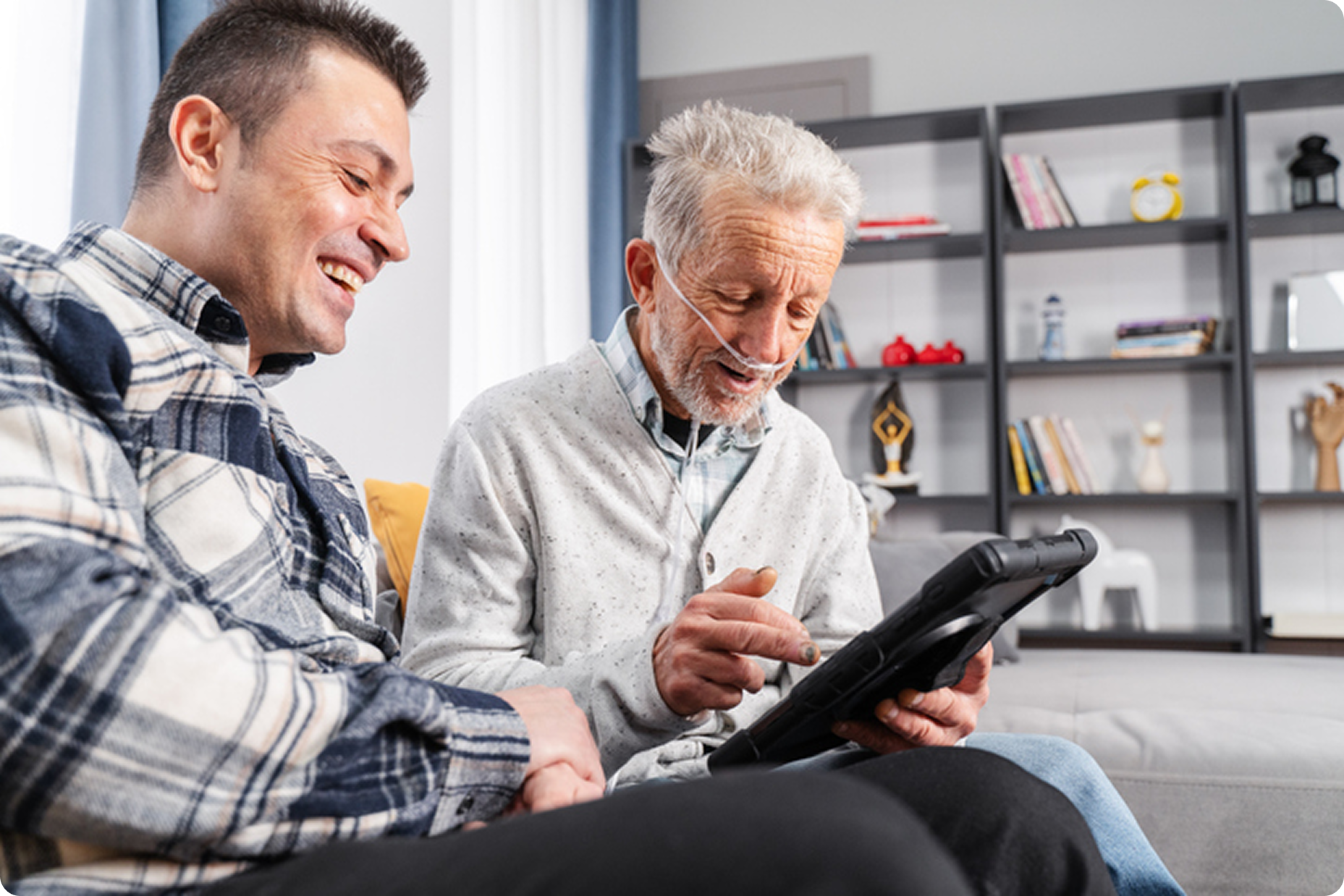 An older man with a nasal cannula smiles while using a tablet alongside a younger man in a cozy living room setting.