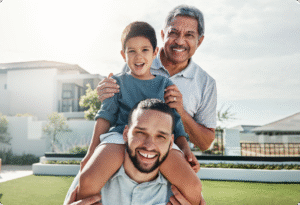 A young boy smiles while being carried on his father's shoulders, with a grandfather standing behind them in a bright outdoor setting.