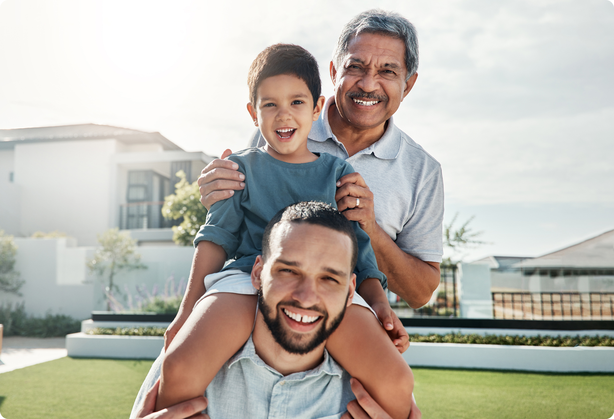 A young boy smiles while being carried on his father's shoulders, with a grandfather standing behind them in a bright outdoor setting.