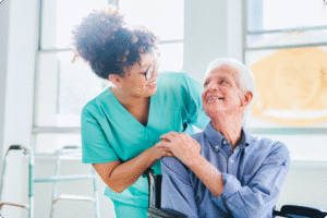 A nurse in scrubs smiles at an elderly man in a wheelchair, sharing a moment of connection in a well-lit room.