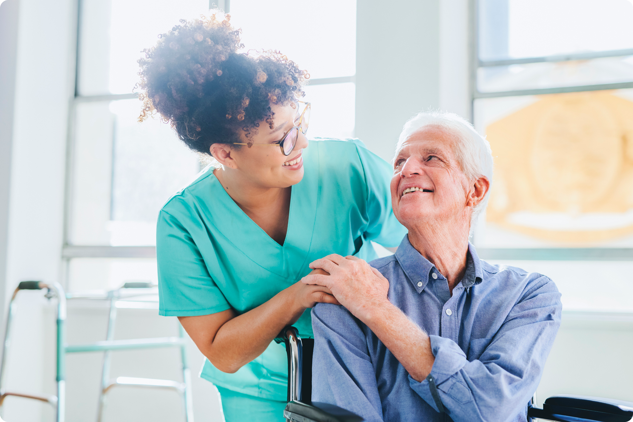 A nurse in scrubs smiles at an elderly man in a wheelchair, sharing a moment of connection in a well-lit room.