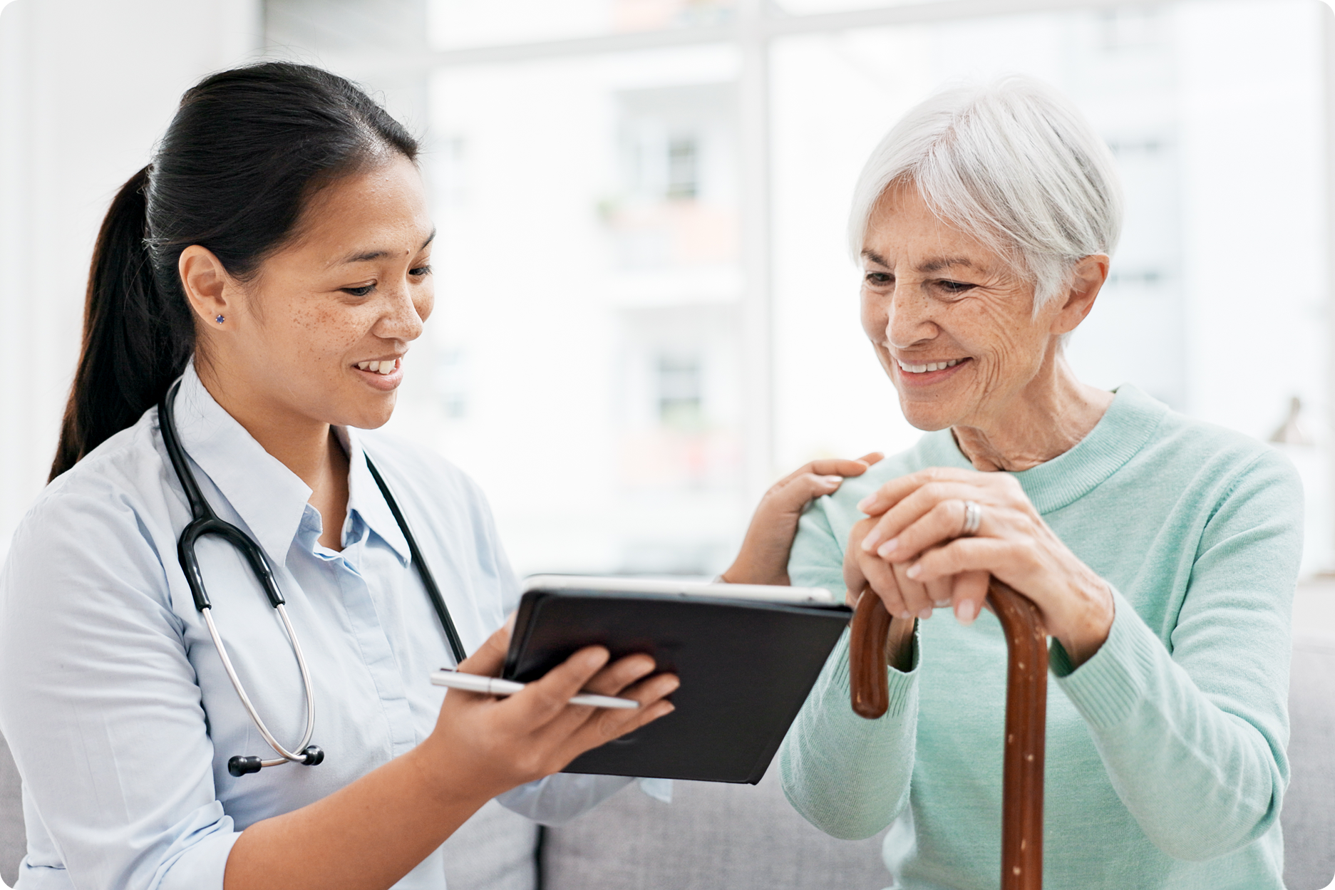 A healthcare professional smiles while discussing information on a tablet with an elderly woman holding a cane.