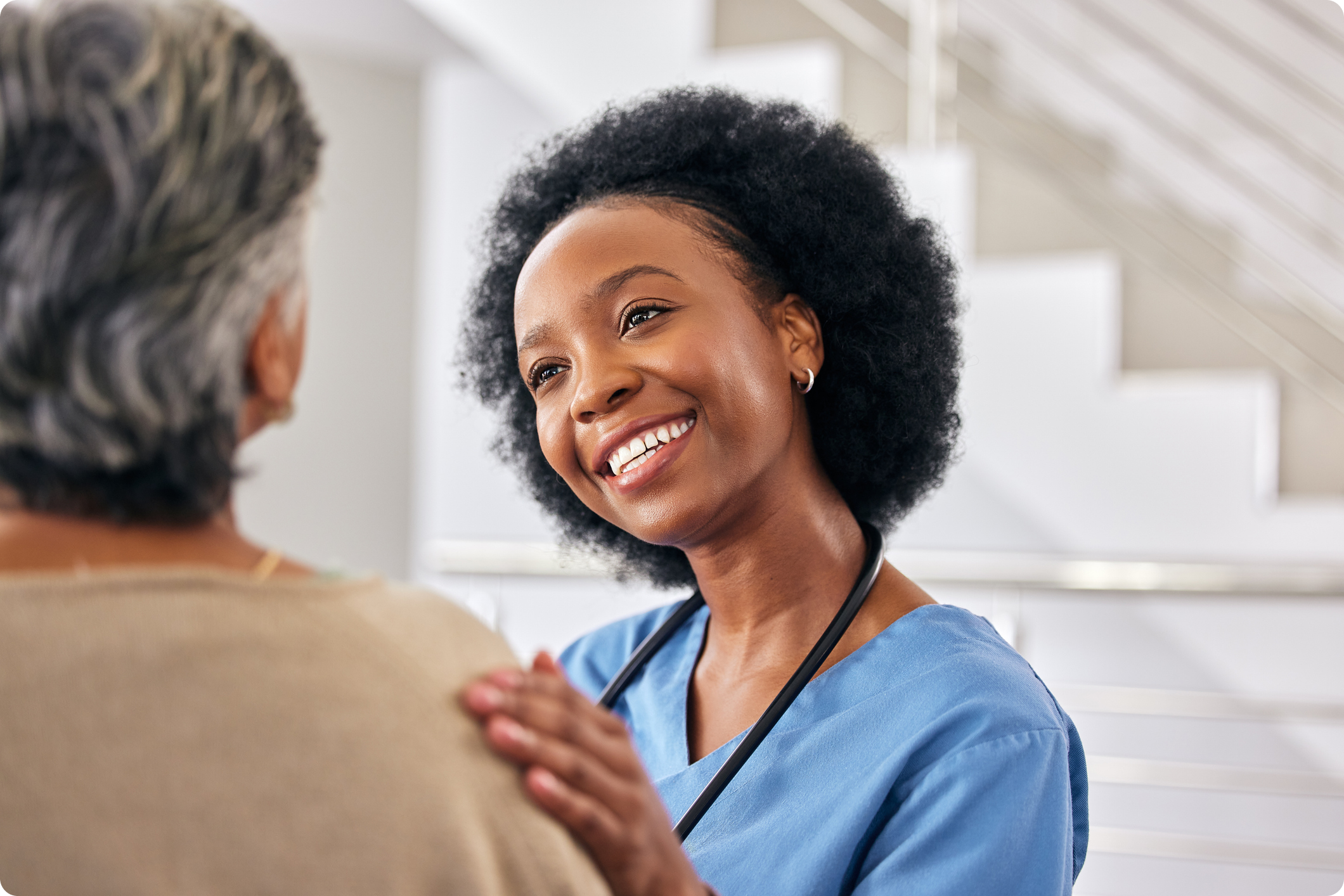 A healthcare professional in scrubs smiles warmly while comforting an elderly patient in a modern medical setting.