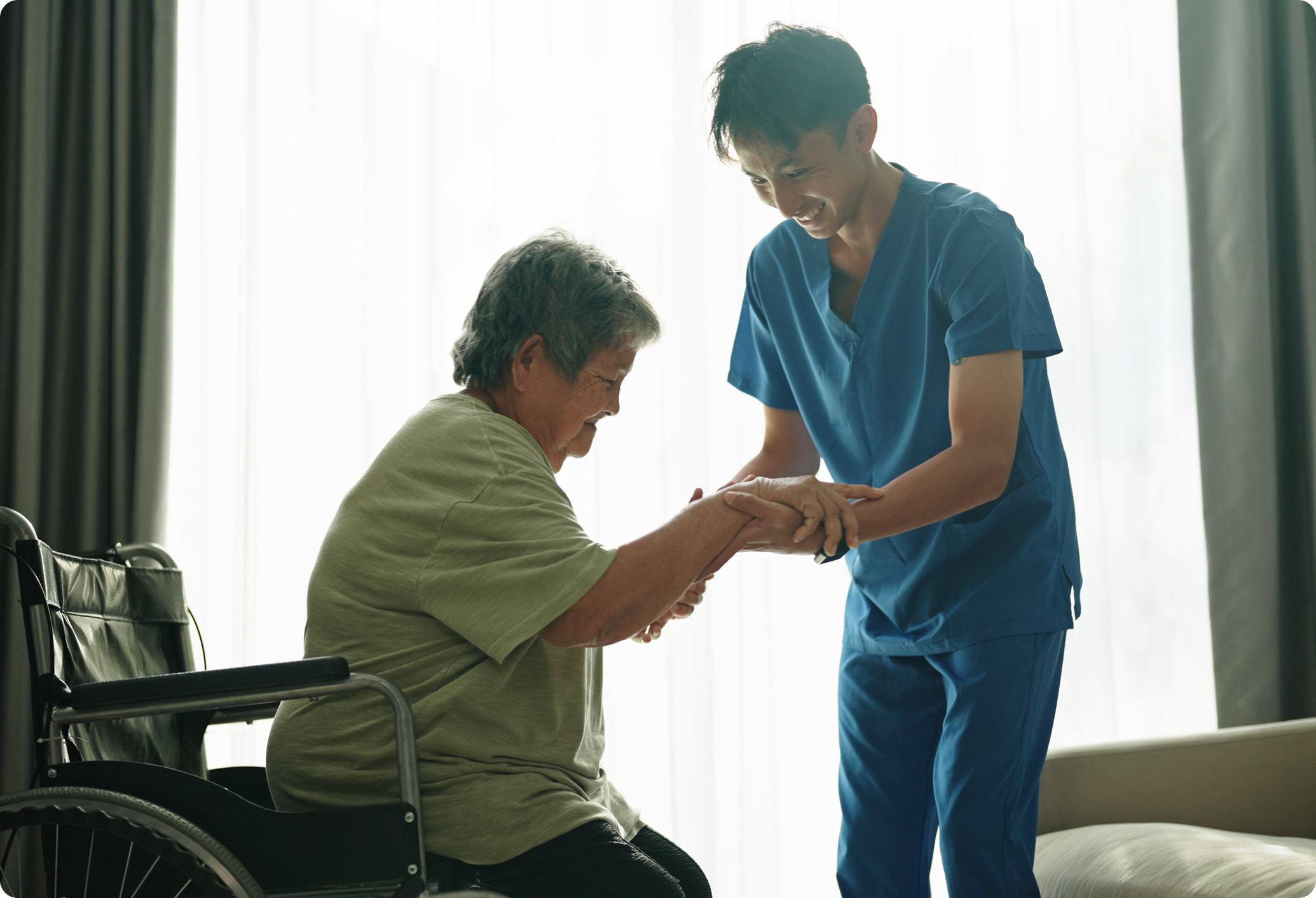 A caregiver assists an elderly woman with mobility, both smiling in a well-lit room with a wheelchair nearby.