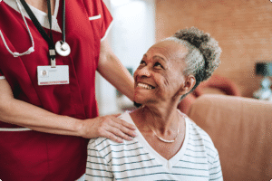 A caregiver in a red uniform supports an elderly woman smiling while seated in a cozy indoor environment.