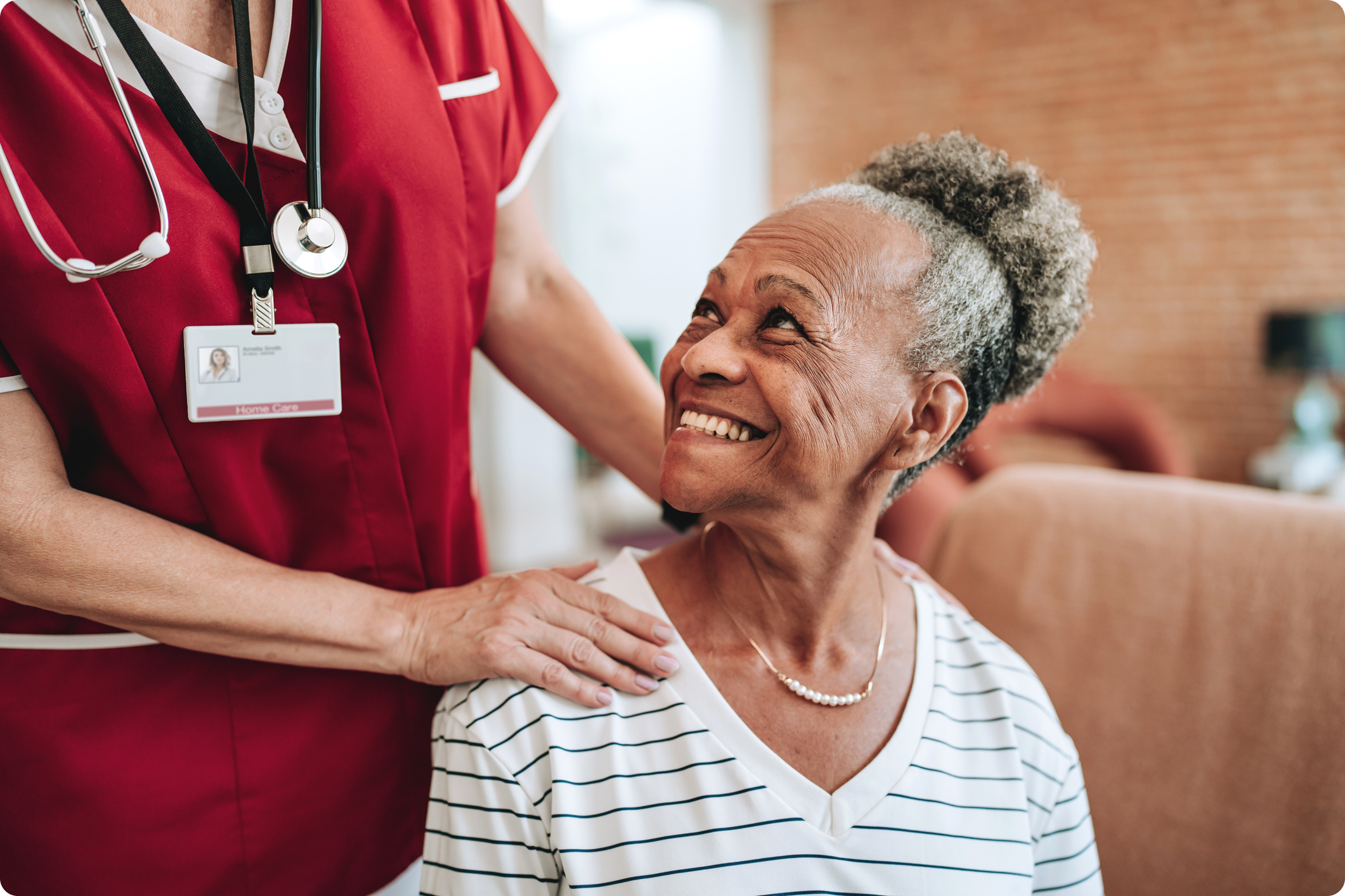 A caregiver in a red uniform supports an elderly woman smiling while seated in a cozy indoor environment.