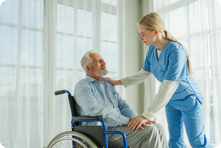 A caring nurse interacts with an elderly man in a wheelchair, both smiling in a bright room with large windows.