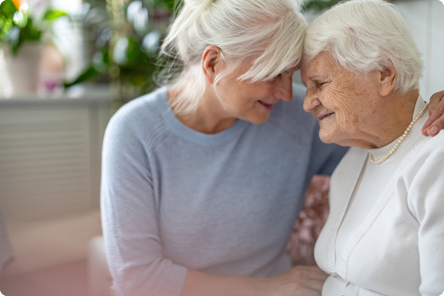 A younger woman with blonde hair smiles affectionately while leaning close to an elderly woman with white hair.