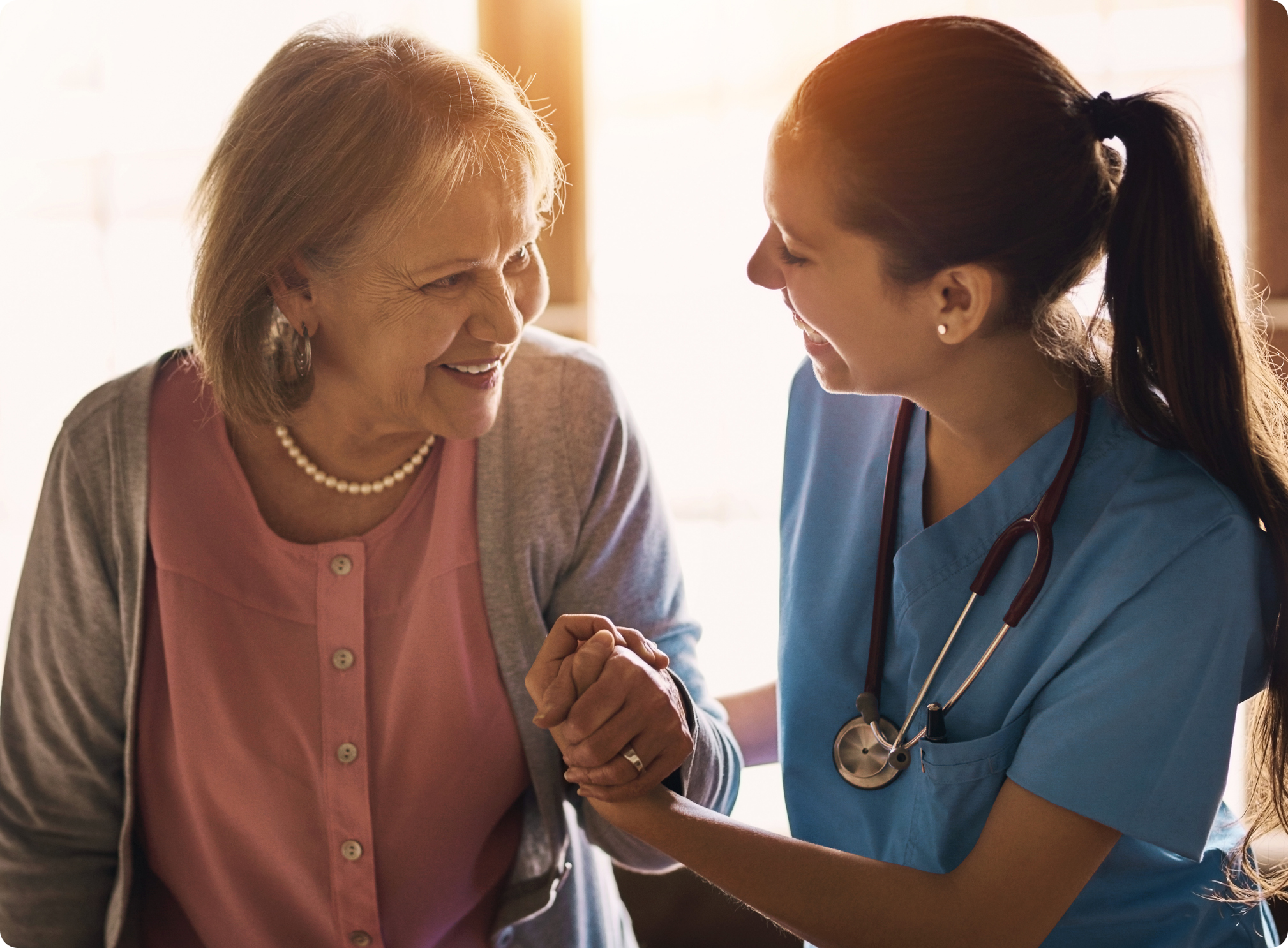 A nurse in blue scrubs engages warmly with an elderly woman, fostering a caring connection in a bright, inviting setting.
