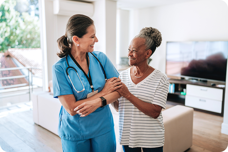 A healthcare professional in blue scrubs interacts warmly with an elderly woman in a well-lit, modern living room.