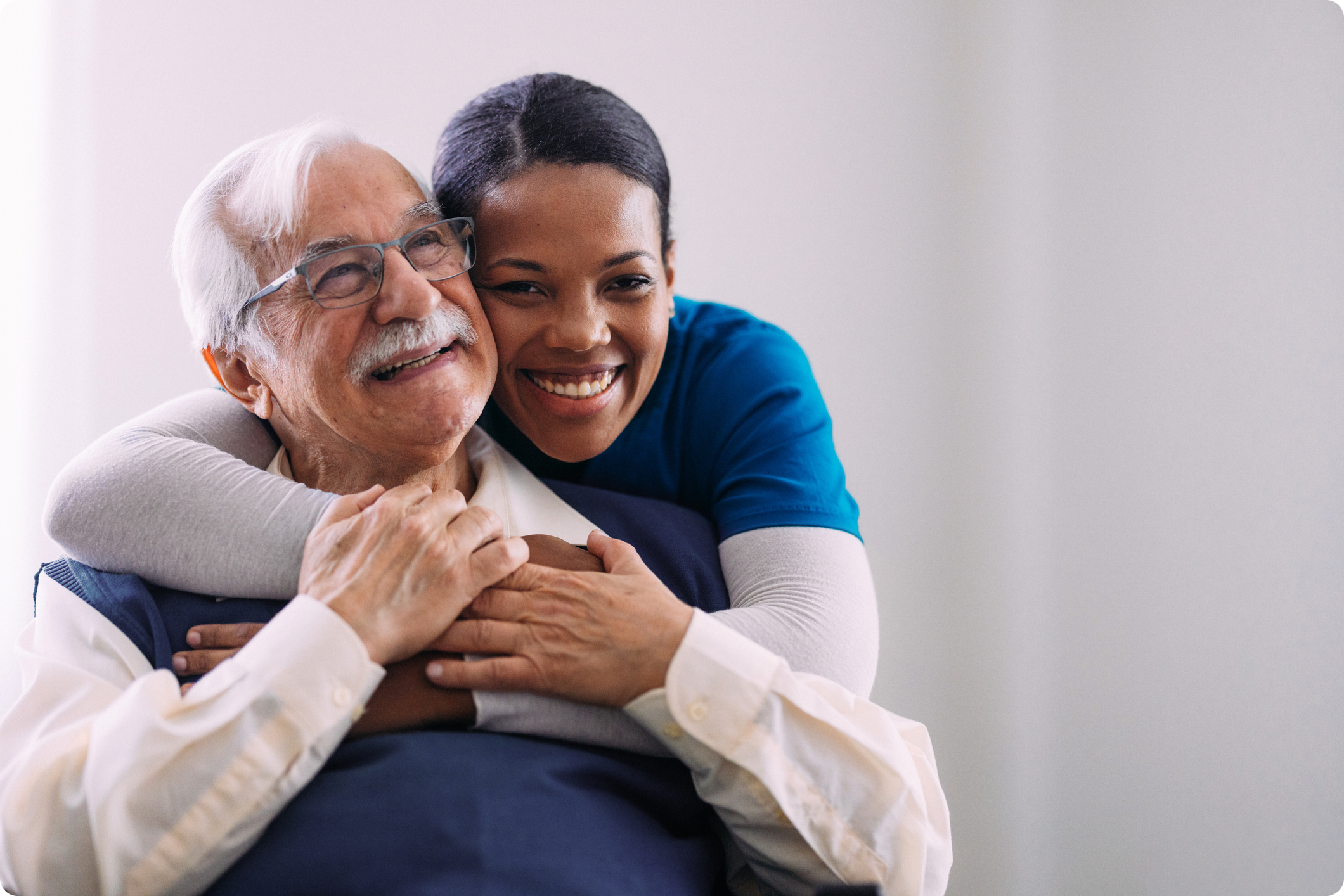 A smiling elderly man is embraced by a woman in scrubs, both conveying warmth and companionship in a bright setting.
