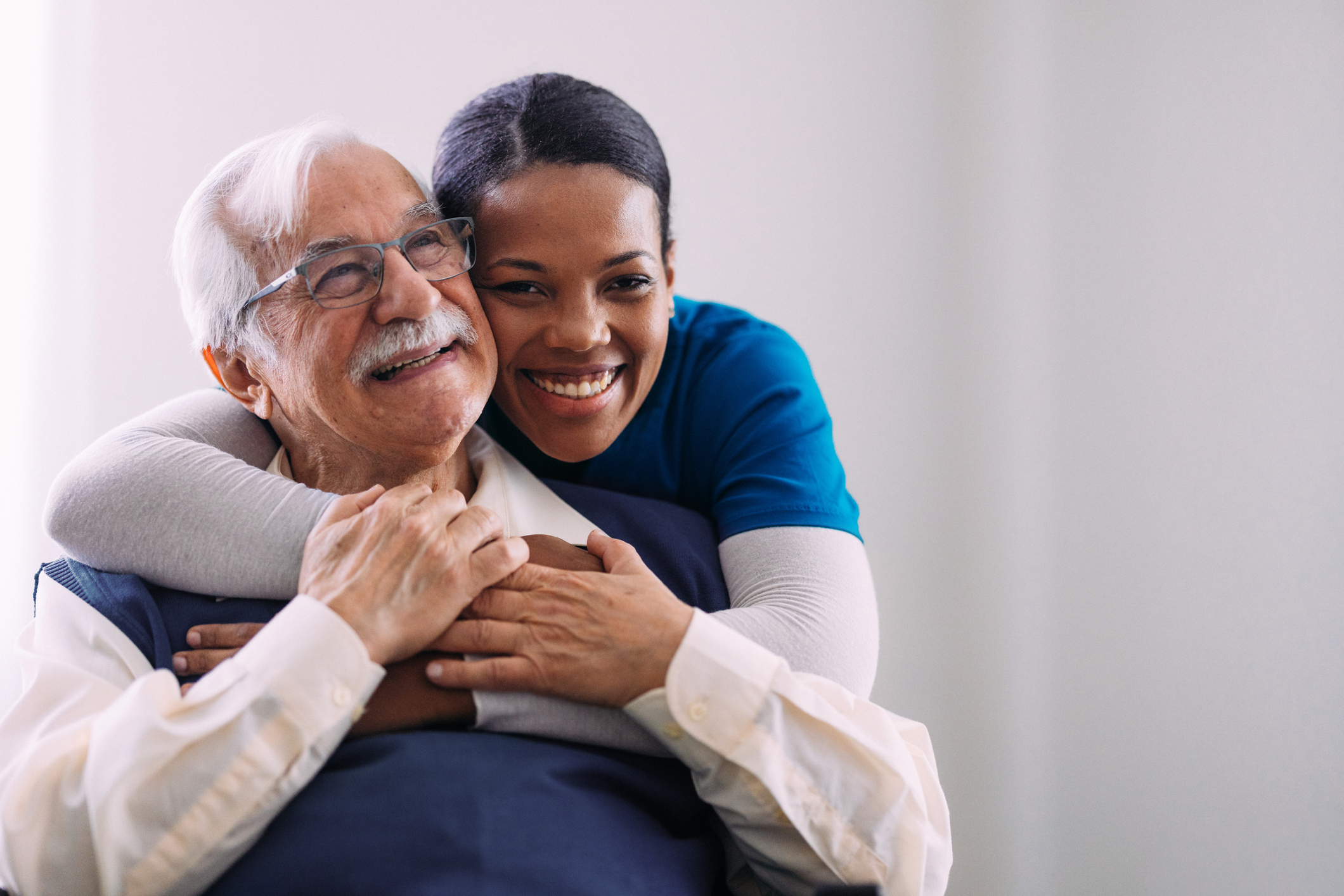 Smiling elderly man in glasses embraces a woman in a blue shirt, highlighting warmth and companionship in a bright setting.