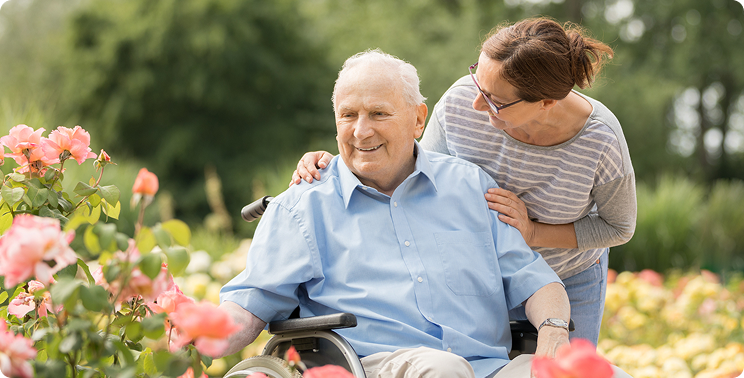 Senior man in a wheelchair smiles as a woman leans over him in a colorful garden, surrounded by blooming flowers.