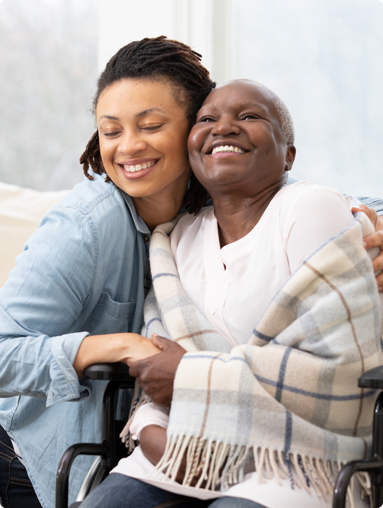 A woman embraces an elderly woman draped in a cozy blanket, both smiling warmly in a bright, inviting interior setting.