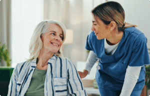 A healthcare professional in blue scrubs is smiling and interacting with an elderly patient in a bright, welcoming environment.