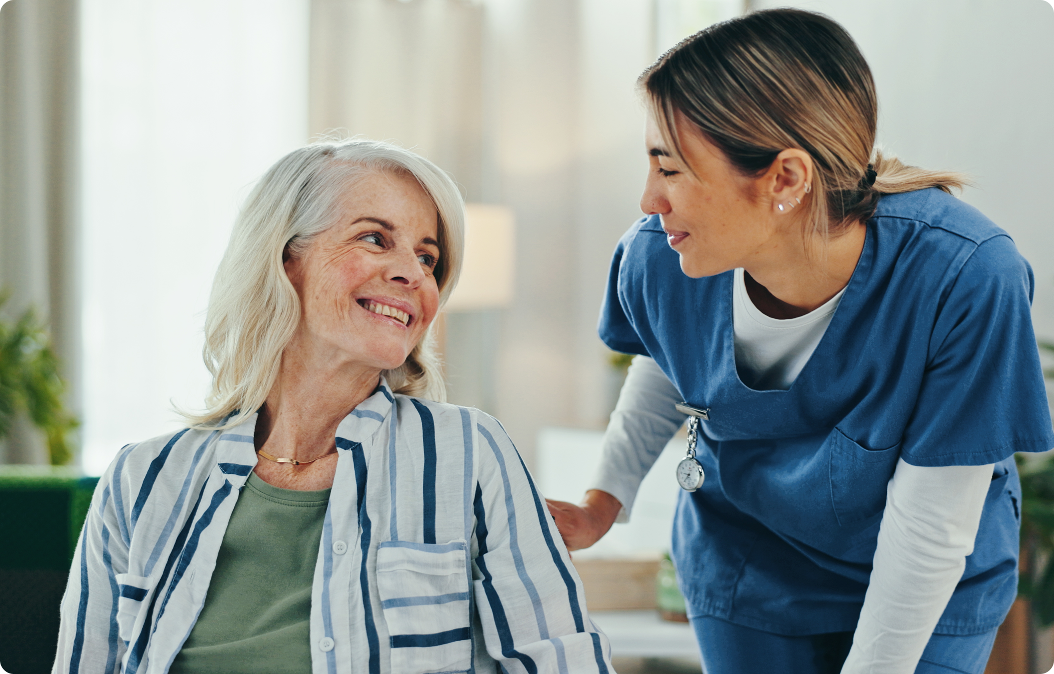 A healthcare professional in blue scrubs is smiling and interacting with an elderly patient in a bright, welcoming environment.