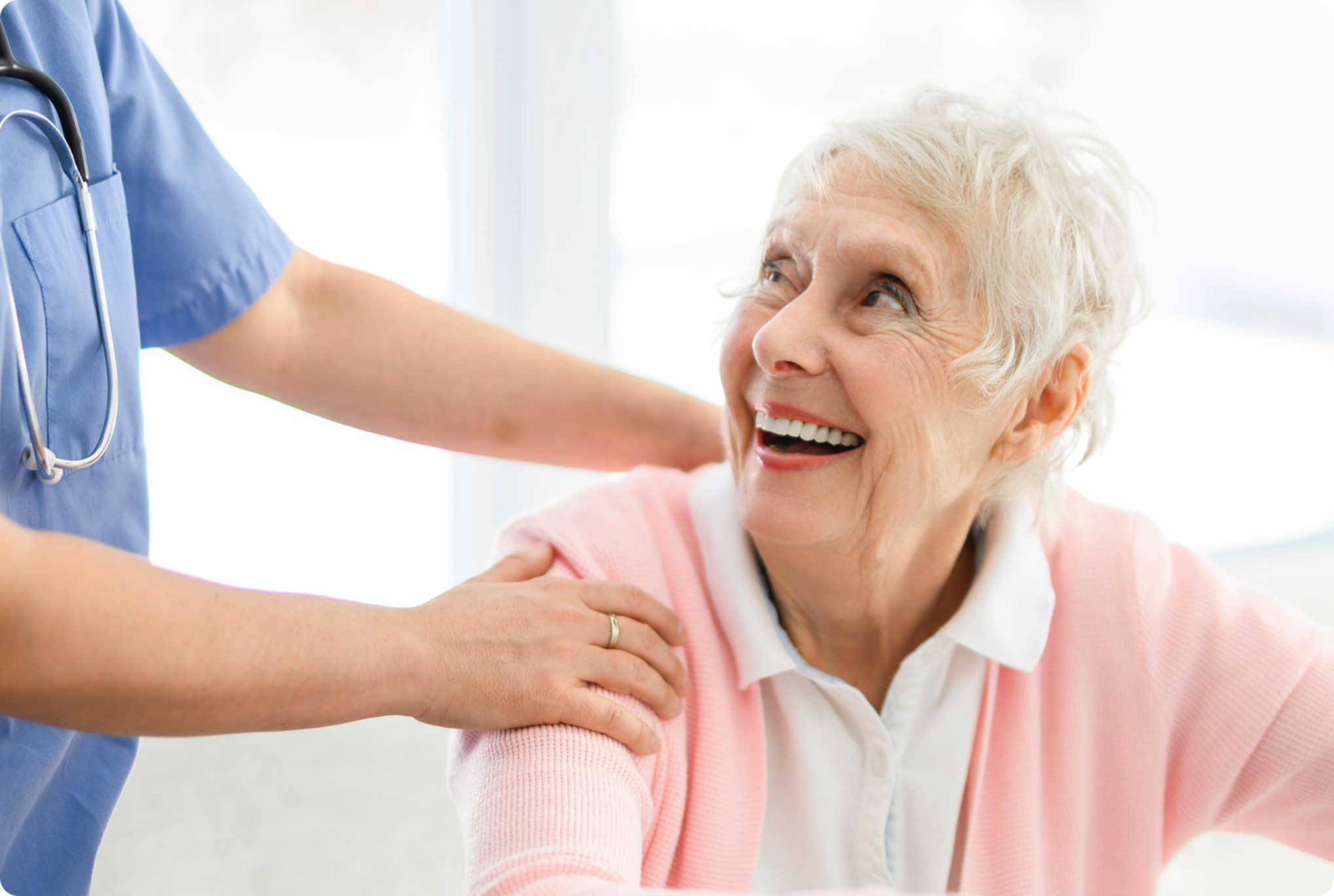 A senior woman smiles brightly while a healthcare professional gently touches her shoulder in a well-lit medical setting.